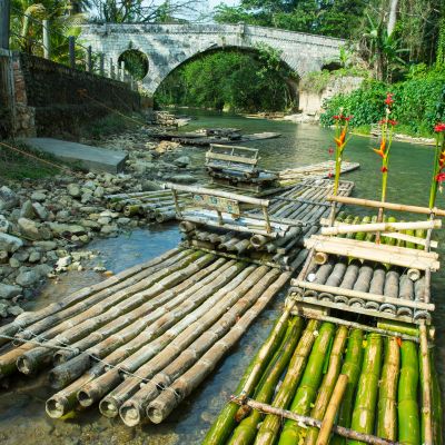 Traditional bamboo rafts lined up along a calm riverbank in Jamaica, with a stone bridge in the background and lush greenery surrounding the scene.