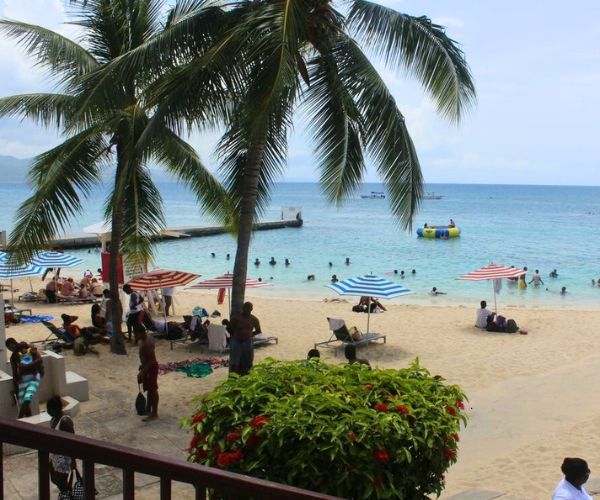 Crowd enjoying a sunny day at Doctors Cave Beach in Jamaica, with palm trees, umbrellas, and people swimming in the turquoise sea.