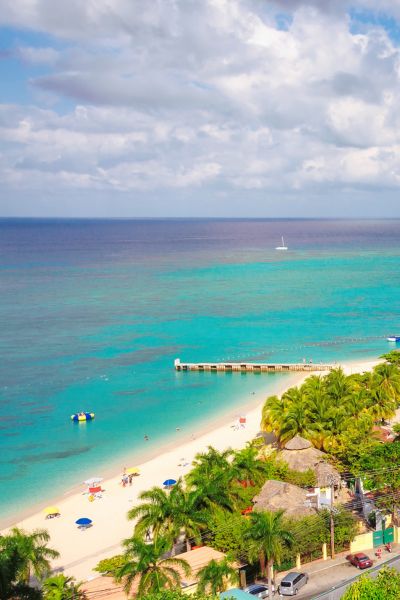 Aerial view of Montego Bay showing a tropical Jamaican beach with turquoise water, palm trees, colorful umbrellas, and a pier stretching into the sea.